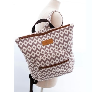 May include: A backpack with a geometric pattern in shades of gray and white. The bag has a front pocket, a top handle, and brown zippers. A small leather tag reads "HANDMADE". The backpack is displayed on a mannequin.