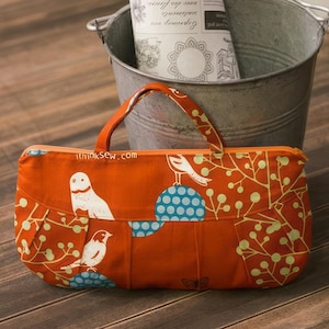 Pode incluir: Uma bolsa de tecido laranja brilhante com zíper, um pássaro branco e um design de bolinhas azuis. A bolsa tem uma alça marrom e o texto "ithinkSew.com" está impresso na frente.