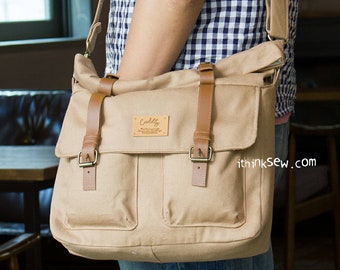 May include: A tan canvas messenger bag with brown leather straps and buckles. The bag has a flap with a brown leather patch that says "Cuddly" on it. The bag is being worn by a person wearing a blue and white gingham shirt.
