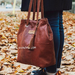 Può includere: Una borsa tote in pelle marrone con un pon pon e il testo "ithinksew.com" sul davanti. La borsa ha una chiusura con cerniera superiore e due manici.