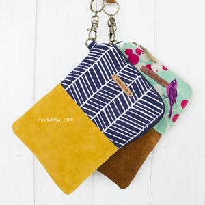 May include: Three small zippered pouches with fabric and suede exteriors. The pouches are hanging from a brown and yellow strap with metal clasps. The pouches have different patterns: one has a blue and white chevron pattern, one has a green and white floral pattern with a bird, and one has a red and white polka dot pattern. The pouches have the text "Limited Edition" and "Hand Made" on them.