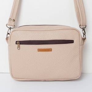 Op de afbeelding: Een lichtbeige schoudertas met een bruine rits en een bruin lederen label met de tekst "HANDMADE".