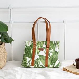 May include: A green and white canvas tote bag with brown leather handles and straps. The bag has a tropical leaf print.