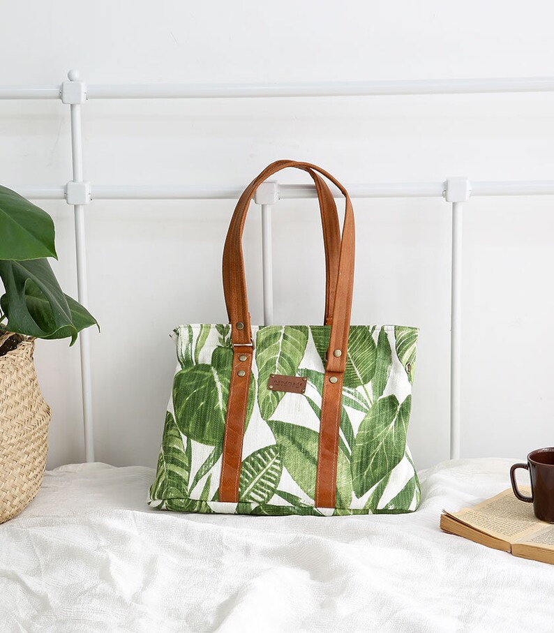 May include: A green and white canvas tote bag with brown leather handles and straps. The bag has a tropical leaf print.