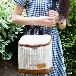 May include: A beige and brown insulated lunch bag with a geometric pattern and a brown leather strap. The bag has a zipper closure and a "handmade" label.