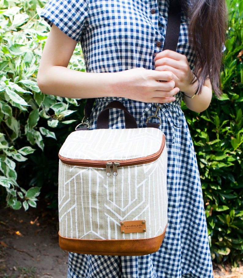May include: A beige and brown insulated lunch bag with a geometric pattern and a brown leather strap. The bag has a zipper closure and a "handmade" label.