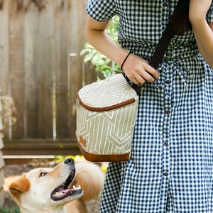 May include: A woman wearing a blue and white gingham dress carries a brown and white insulated lunch bag with a shoulder strap. A light brown dog looks up at the bag.