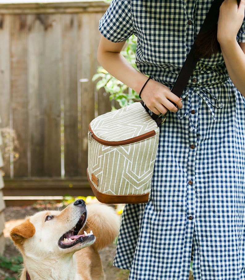 May include: A woman wearing a blue and white gingham dress carries a brown and white insulated lunch bag with a shoulder strap. A light brown dog looks up at the bag.