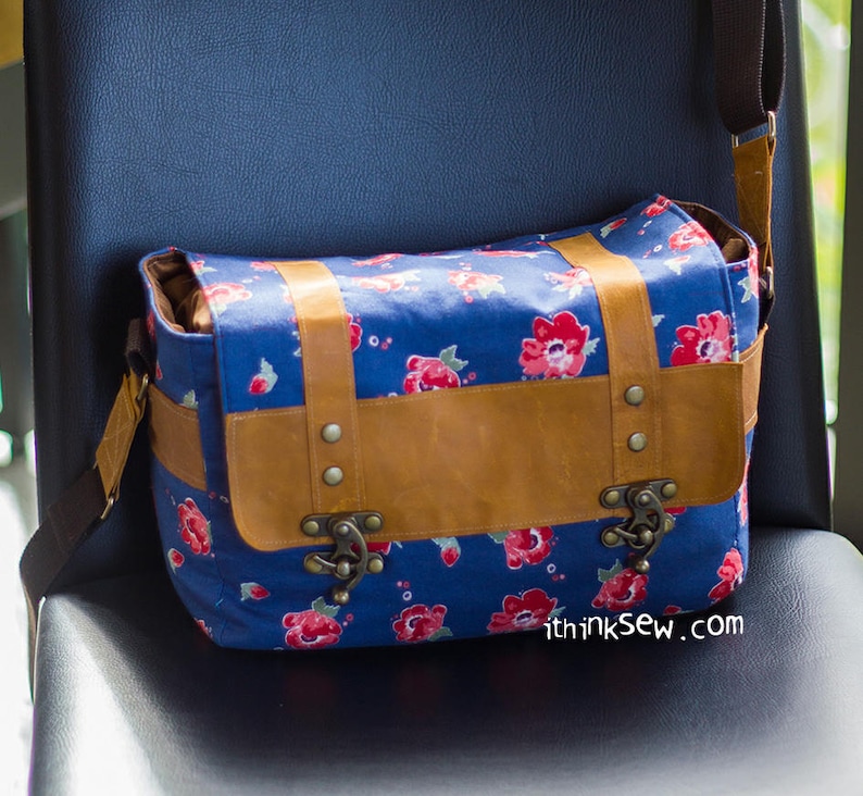 Pu&ograve; includere: Una borsa a tracolla in tela blu con una patta in pelle marrone e fibbie metalliche. La borsa ha una stampa floreale con fiori rossi e punti bianchi. La borsa ha una tracolla e il testo "thinksew.com" &egrave; stampato sul fondo.