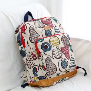 May include: A white canvas backpack with a red zipper and a brown leather bottom. The backpack has a colorful pear pattern with blue, green, red, and yellow accents. The text "handmade" is visible on the front of the bag.