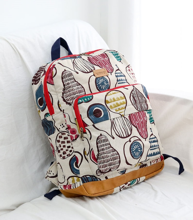 May include: A white canvas backpack with a red zipper and a brown leather bottom. The backpack has a colorful pear pattern with blue, green, red, and yellow accents. The text "handmade" is visible on the front of the bag.