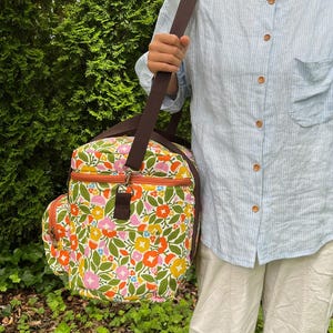 May include: A floral print bag with a brown shoulder strap and a smaller side pocket. The bag features a colorful pattern of flowers in orange, pink, yellow, and blue on a white background. The bag has an orange zip and brown trim.