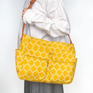 Puede incluir: Una bolsa de lona amarilla con un patrón geométrico blanco y varios bolsillos. La bolsa tiene una correa de cuero marrón.