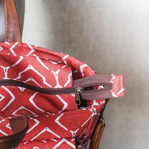K&ouml;nnte beinhalten: Nahaufnahme einer roten Canvas-Tragetasche mit einem wei&szlig;en geometrischen Muster. Die Tasche hat einen braunen Ledergriff und einen braunen Rei&szlig;verschluss mit einem goldenen Zipper.