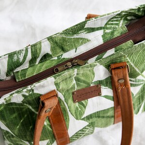 May include: A white canvas tote bag with a green tropical leaf print. The bag has brown leather handles and a brown zipper closure. The bag has a brown leather tag that says "handmade with love".