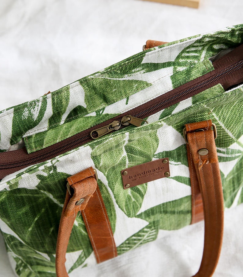 May include: A white canvas tote bag with a green tropical leaf print. The bag has brown leather handles and a brown zipper closure. The bag has a brown leather tag that says "handmade with love".