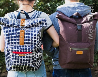 May include: Two people wearing backpacks. The backpack on the left is blue and white with a red zipper. The backpack on the right is brown with a black buckle and a green zipper. Both backpacks have a roll-top closure.