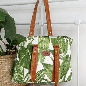 May include: A white canvas tote bag with a green tropical leaf print and brown leather straps and handles. The bag has a brown leather tag that says "handmade".