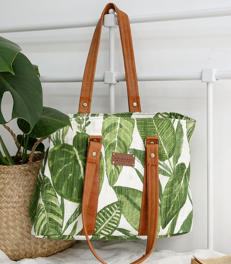 May include: A white canvas tote bag with a green tropical leaf print and brown leather straps and handles. The bag has a brown leather tag that says "handmade".