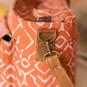 K&ouml;nnte beinhalten: Nahaufnahme einer leuchtend orangefarbenen Canvas-Tragetasche mit einem wei&szlig;en geometrischen Muster. Die Tasche hat einen braunen Lederriemen mit einem Metallverschluss.