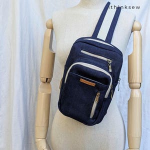 May include: A dark blue denim sling backpack featuring a single wide off-white shoulder strap, multiple zippered compartments with off-white trim, and a small rectangular brown leather tag labeled "HANDMADE" on the front pocket.