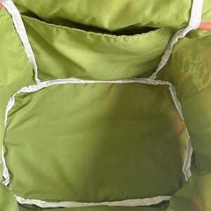 May include: Close-up of the interior of a green tote bag. The bag features a large main compartment and a smaller pocket, both lined with a lighter green fabric. White trim outlines the edges of the pockets and the bag's opening.