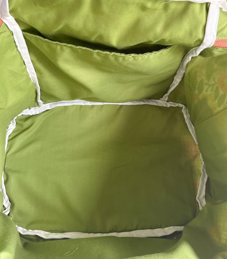 May include: Close-up of the interior of a green tote bag. The bag features a large main compartment and a smaller pocket, both lined with a lighter green fabric. White trim outlines the edges of the pockets and the bag's opening.