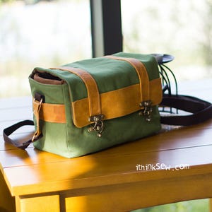Pu&ograve; includere: Una borsa per fotocamera in tela verde con dettagli in pelle marrone e chiusure in metallo. La borsa ha una tracolla ed &egrave; appoggiata su un tavolo di legno.