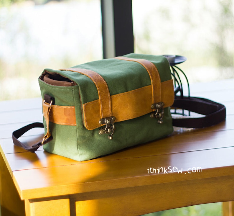 Pu&ograve; includere: Una borsa per fotocamera in tela verde con dettagli in pelle marrone e chiusure in metallo. La borsa ha una tracolla ed &egrave; appoggiata su un tavolo di legno.