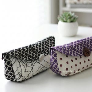 Könnte beinhalten: Zwei Stoffetuis mit Knopfverschlüssen. Ein Etui ist schwarz-weiß mit einem geometrischen Muster und einem weißen Blattdesign. Das andere ist lila und weiß mit einem Tupfenmuster. Beide Etuis sind rechteckig.