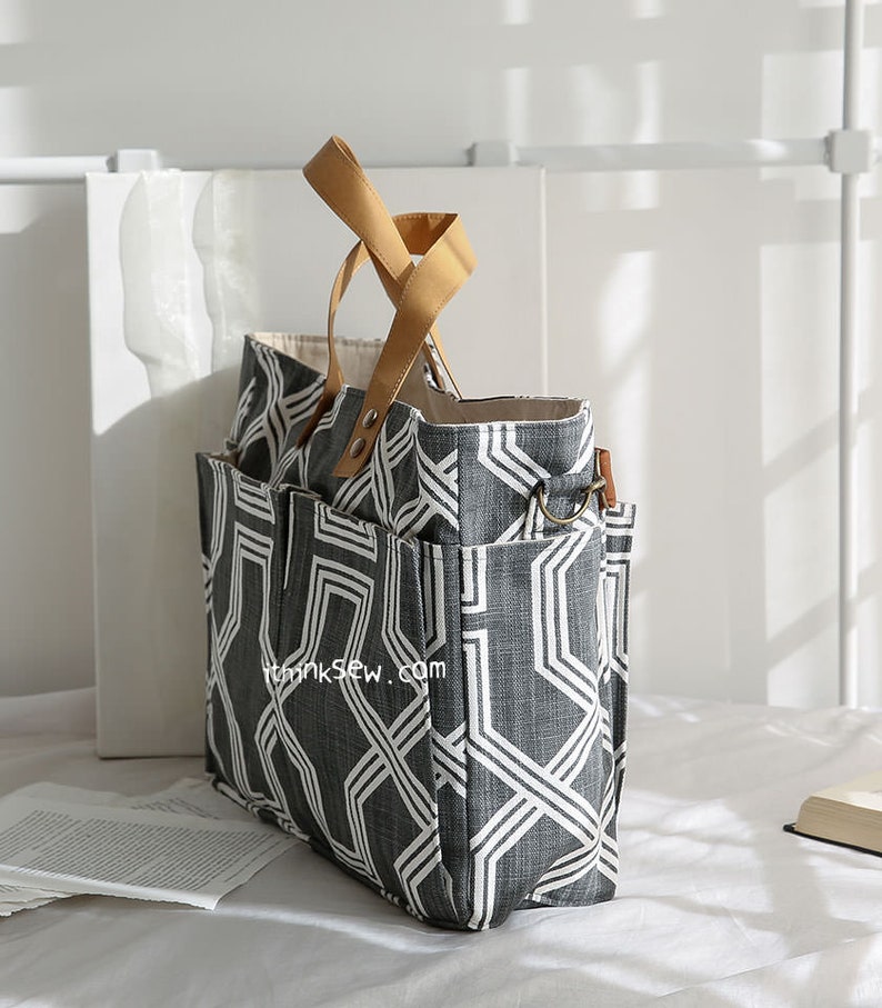 May include: A large gray and white geometric patterned tote bag with brown leather handles. The bag has multiple pockets and a zipper closure.