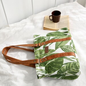 May include: A white canvas tote bag with a green tropical leaf print and brown leather straps and accents. The bag has a brown leather label with the text "Handmade with Love" on it.