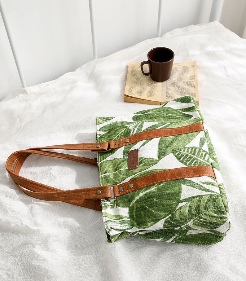 May include: A white canvas tote bag with a green tropical leaf print and brown leather straps and accents. The bag has a brown leather label with the text "Handmade with Love" on it.