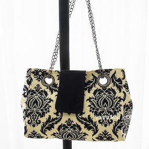 Może przedstawiać: Prostokątna torebka z czarno-kremowym wzorem damasceńskim. Torba posiada czarne zapięcie na klapę i jest zawieszona na srebrnym łańcuszku. Torba ma duże srebrne przelotki i czarny uchwyt.