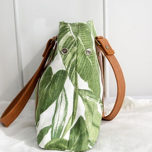 May include: A green and white tote bag with a leafy pattern. The bag has brown leather straps and metal accents.