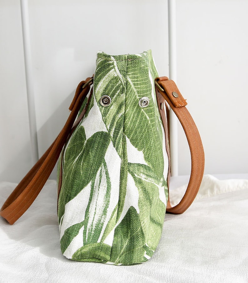May include: A green and white tote bag with a leafy pattern. The bag has brown leather straps and metal accents.