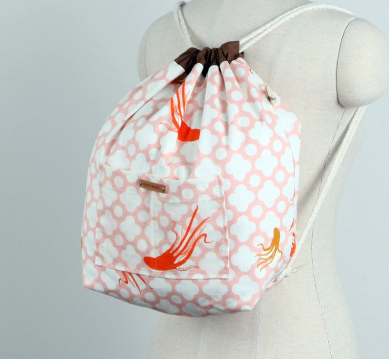May include: A drawstring backpack featuring a white and light pink quatrefoil pattern with orange octopus illustrations. The bag has a brown fabric top closure, white rope straps, and a front pocket with a small rectangular tag labelled HAND MADE.