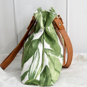 May include: A white canvas tote bag with a green tropical leaf print and brown leather straps.