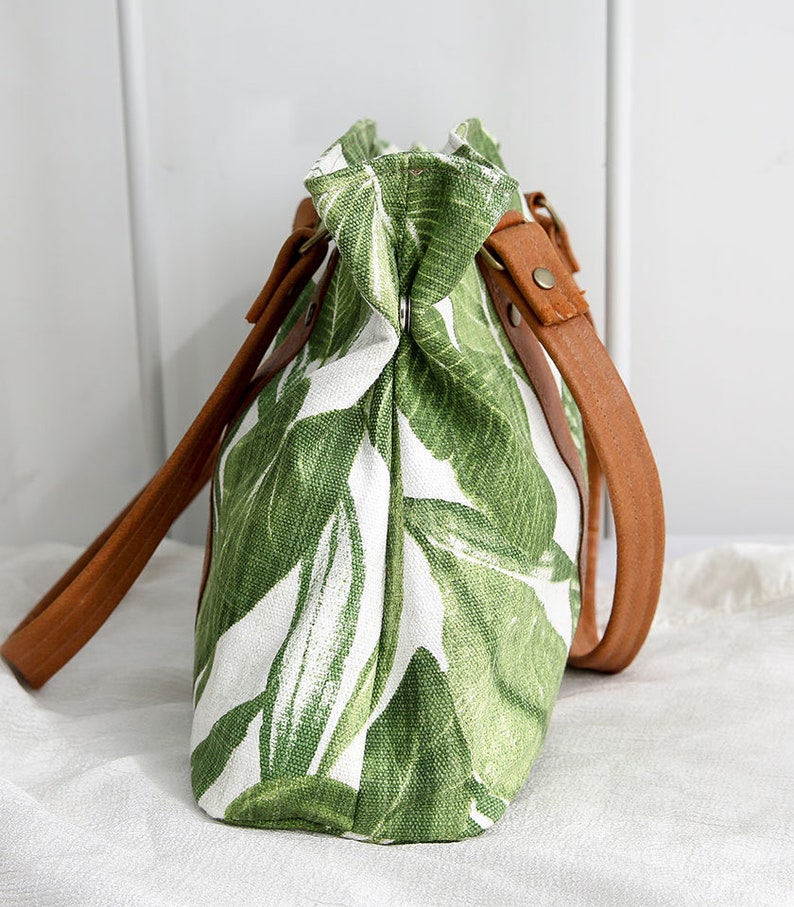 May include: A white canvas tote bag with a green tropical leaf print and brown leather straps.