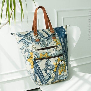 Puede incluir: Un bolso tote grande de lona blanca con un estampado floral azul y amarillo. El bolso tiene asas de cuero marrón y un bolsillo con cremallera en la parte delantera. El bolso está sentado en una silla blanca.
