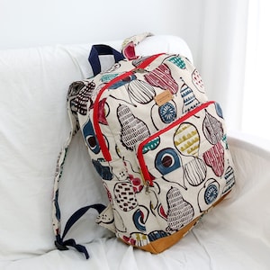 May include: A white canvas backpack with a red zipper and a brown leather bottom. The backpack is covered in a colorful pattern of pears and other fruits. The text "handmade" is printed on a brown leather tag.