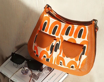 Puede incluir: Bolso de hombro con estampado naranja y blanco, ribete de cuero marrón y dos bolsillos delanteros. El bolso está junto a un libro, gafas de sol y joyas. El diseño del bolso presenta detalles en negro.