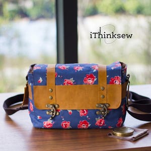 Blue floral fabric and brown leather vintage camera bag with antique brass clasps sitting on a wooden table. A handmade padded DSLR crossbody bag sewing pattern PDF with a lens cap and cleaning cloth nearby.