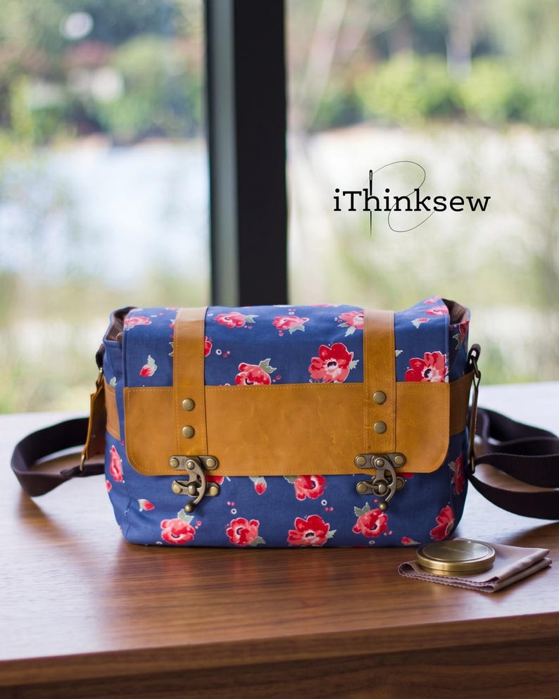 Blue floral fabric and brown leather vintage camera bag with antique brass clasps sitting on a wooden table. A handmade padded DSLR crossbody bag sewing pattern PDF with a lens cap and cleaning cloth nearby.