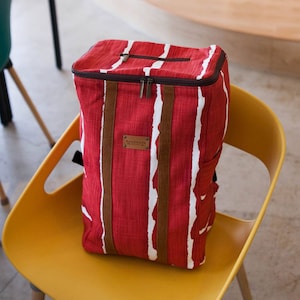 Può includere: Zaino in tela a righe rosse e bianche con dettagli in pelle marrone e chiusura a zip. Lo zaino ha un'etichetta "fatto a mano" sul davanti.