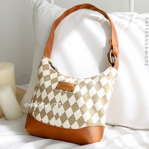 May include: A brown and white checkered shoulder bag with a brown leather bottom and a brown leather strap. The bag has a metal ring on the strap and a small leather tag that says "handmade". The bag is sitting on a white bedspread.