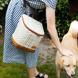 May include: A woman in a blue and white gingham dress is petting a light brown dog. She is carrying a beige and brown insulated lunch bag with a white geometric pattern. The bag has a zipper closure and a brown leather trim.