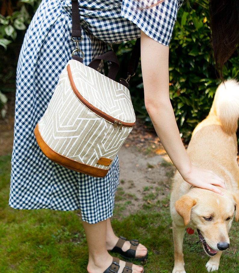 May include: A woman in a blue and white gingham dress is petting a light brown dog. She is carrying a beige and brown insulated lunch bag with a white geometric pattern. The bag has a zipper closure and a brown leather trim.