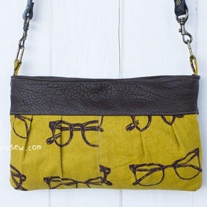 May include: A mustard yellow fabric crossbody bag with a brown leather flap and a black and white eyeglasses print. The bag has a long adjustable strap with a silver clasp.