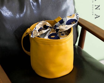 May include: A mustard yellow leather bucket bag with a fabric lining featuring a floral pattern in shades of white, yellow, and blue. The bag has a single strap and is sitting on a black leather chair.